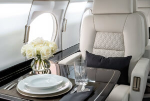 Luxury private jet interior featuring a cream leather seat with quilted stitching, fine dining table setup, crystal glassware, and a bouquet of white roses by the window.