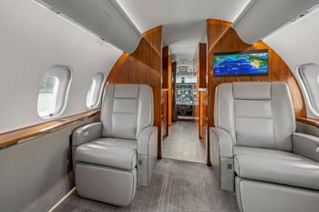 Forward cabin view of a luxury private jet in Sydney with grey leather seats, polished wood paneling, and a visible cockpit through the open divider