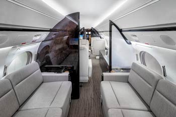 Spacious private jet lounge cabin with dual gray sofas, glossy rear bulkhead, and moving map display, photographed at Van Nuys.
