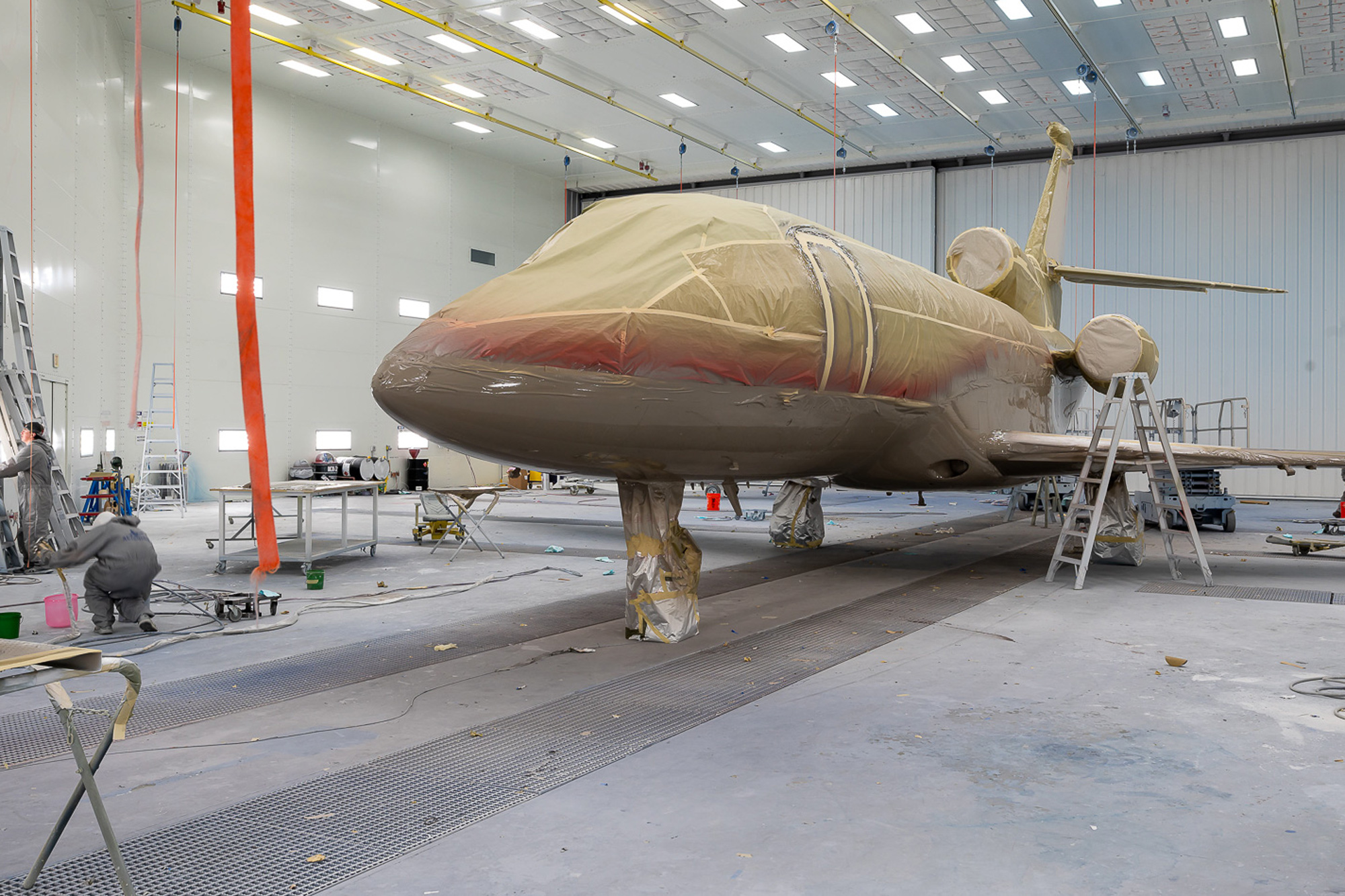 Private jet fully masked for paint in a Wichita hangar, with workers and red guide tape visible on the fuselage.