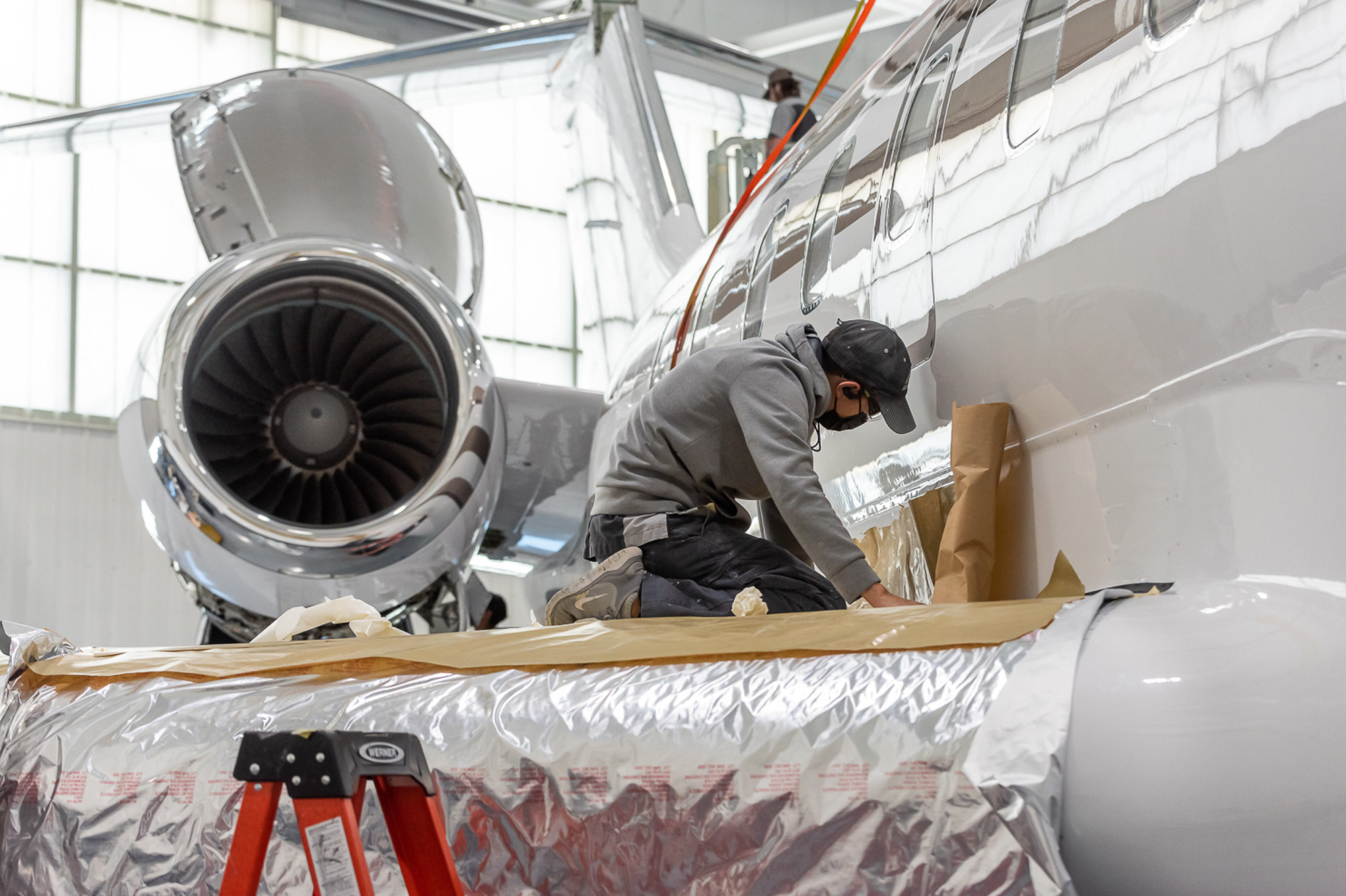 Technician on ladder preps a private jet for paint in a Dallas hangar, working near the engine nacelle with masking in place.