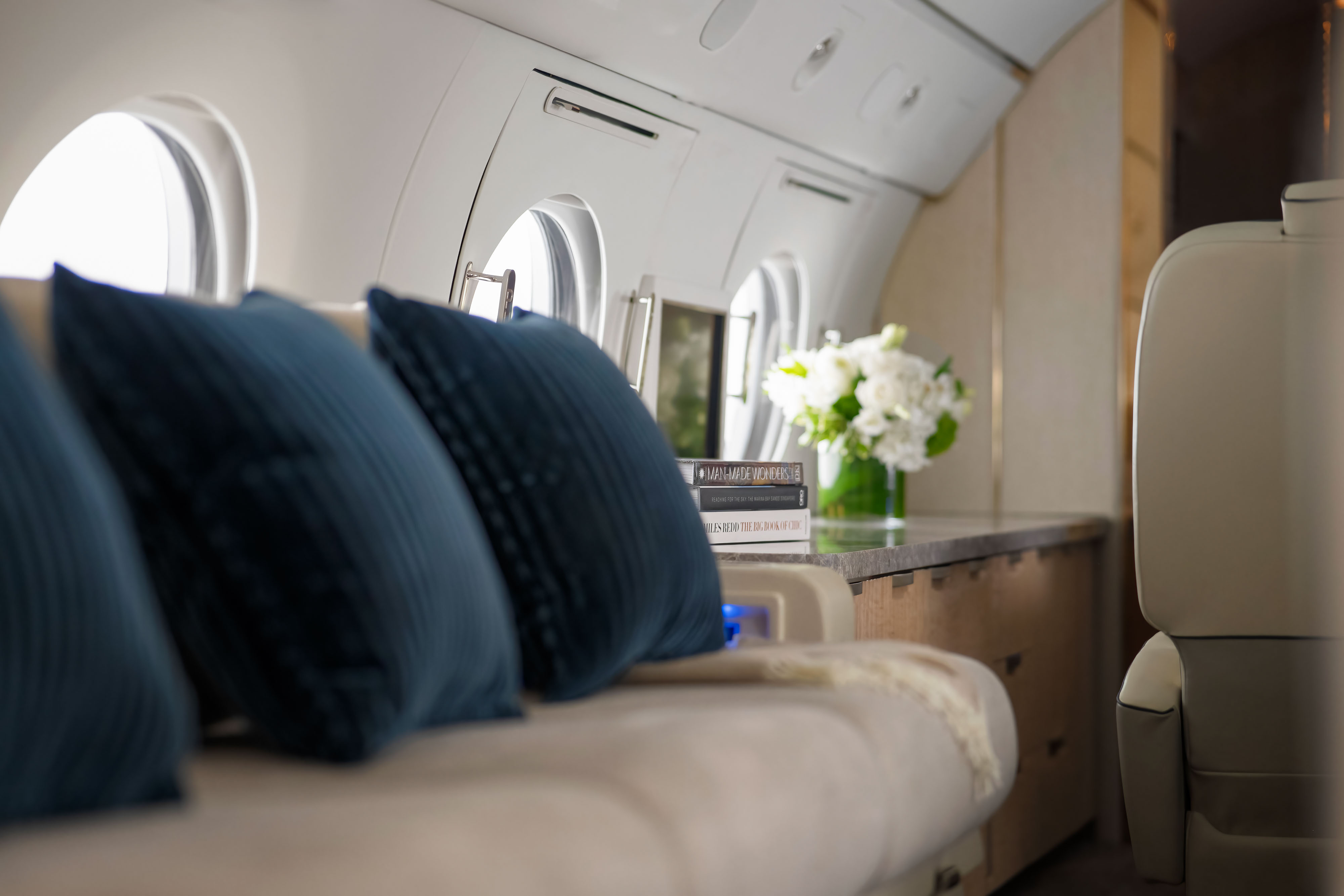 Close-up of a luxurious private jet interior showing a beige sofa with deep blue pillows, a countertop with stacked design books, and a vase of white flowers under softly lit cabin windows.